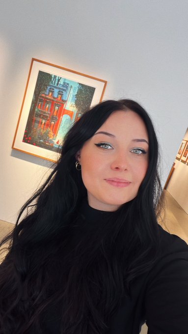 A selfie of a woman with long dark hair wearing a black top, posing in front of a framed colorful painting of a building with a red facade.