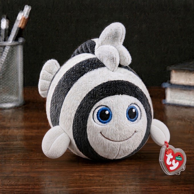 A round, black and white striped plush toy with a smiling face and big blue eyes sits on a wooden surface, featuring a 'ty' heart-shaped tag.