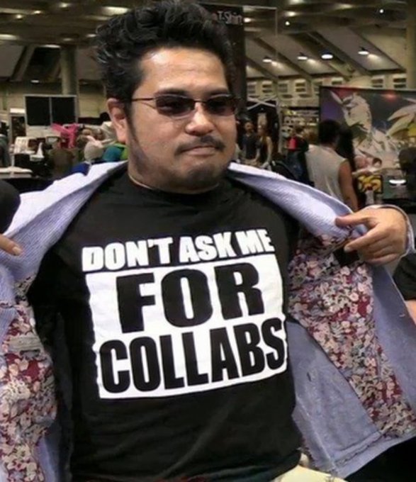 A man wearing glasses and a black t-shirt that reads 'DON'T ASK ME FOR COLLABS' pulls open a floral-patterned jacket at what appears to be a convention.