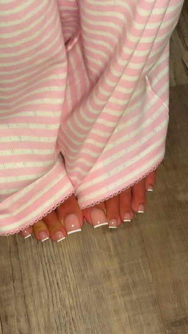 A close-up of a person's feet with a French manicure peeking out from under a pink and white striped knit dress on a wooden floor.