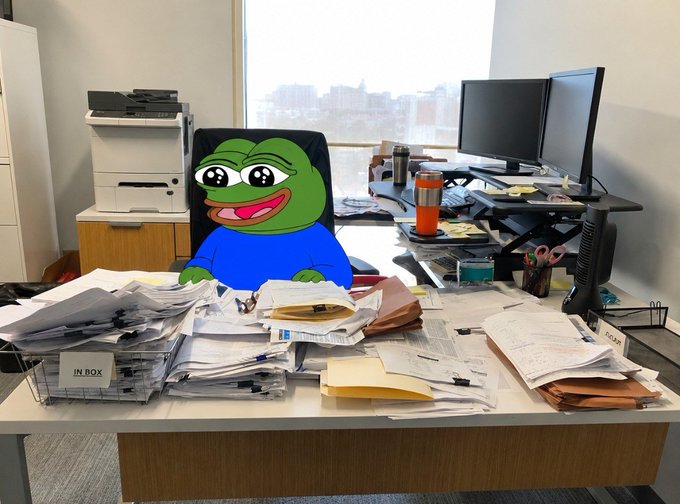 The caption should read: "A Pepe the Frog character sits behind a cluttered desk overflowing with papers, an 'IN BOX' tray stuffed with mail, and office supplies, with a printer and dual monitors in the background."

The image ties into the post's joke by showing the chaotic, paper-strewn workspace that supposedly pays for $peepo's "green candles"—a trading slang reference to profitable price movements—playing on the meme that traders prioritize charts over their actual jobs.