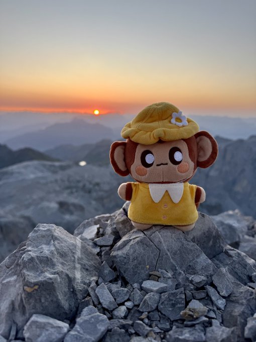 A yellow monkey plush toy sits on a rocky mountain peak with a sunrise visible in the background over a sea of clouds.