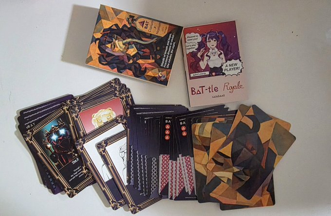 A tabletop card game called "BAT-tle Royale (Calabasas)" is displayed on a white surface, featuring fanned-out stacks of ornately framed cards in dark and orange tones, alongside two character cards—one with an "EXTRAVAGANZA" comic-style badge and another featuring a woman labeled "A NEW PLAYER!"—showcasing the physical release of the community-designed game.