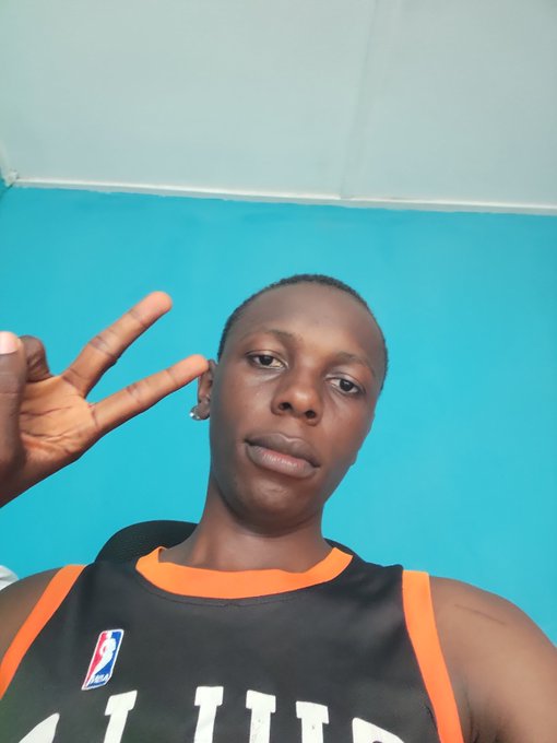 A selfie of a person wearing a black and orange jersey, making a peace sign against a blue and white background.
