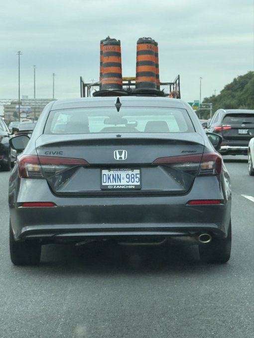 A gray Honda Civic sedan on a highway carrying two orange and black traffic cones strapped to its roof rack, with Ontario license plate 'DKNN-985'.