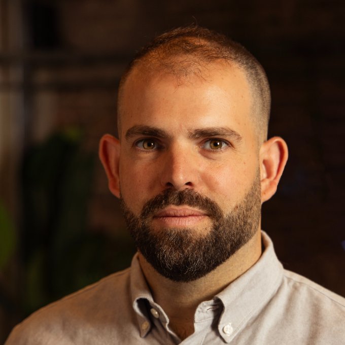A close-up portrait of a man with a short buzz cut, a full beard, and green eyes, wearing a light grey button-up shirt.
