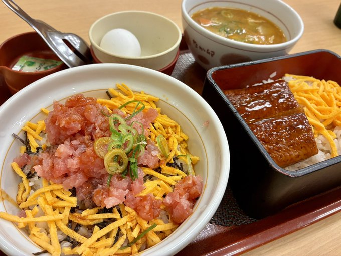 A Japanese meal featuring a bowl topped with minced tuna, shredded egg, and green onions, accompanied by a side of grilled eel over rice, miso soup, a boiled egg, and a small dish of pickled vegetables.