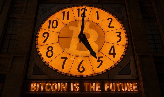 A large illuminated clock displays the time as approximately 12:05, featuring a Bitcoin "₿" symbol at its center and the bold text "BITCOIN IS THE FUTURE" glowing beneath it, visually reinforcing the post's theme of Bitcoin's forward-looking significance.