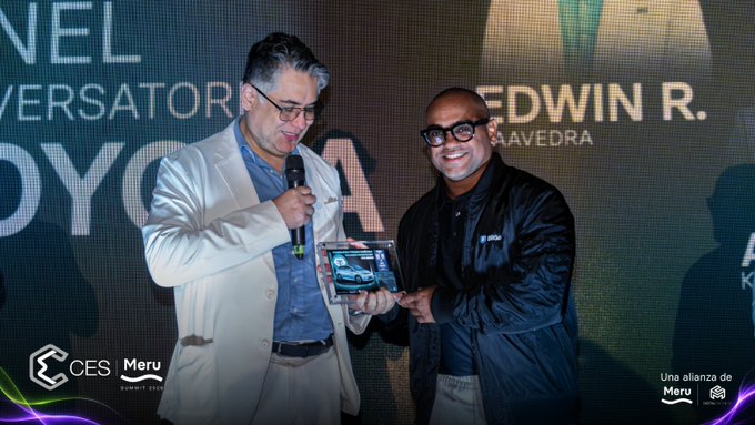 Two men on stage at the Bolivia Crypto Experience Summit, with one in a cream blazer speaking into a microphone while handing a framed car photo to a smiling man in a black jacket, in front of a backdrop reading "EDWIN R. SAAVEDRA" — relating to the post by capturing the moment Alvaro Olivares of BitGo and panelists from Toyota, Towerbank, and Tether celebrated the world's first car sale using USDT.