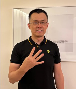 A man in a black polo shirt with a yellow logo smiles and holds up three fingers in front of a framed artwork on a white wall.