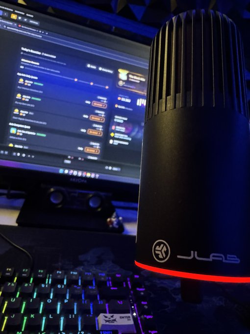 The image features a computer monitor with a keyboard and mouse nearby. The monitor displays a website or webpage, possibly related to gaming or streaming content. In front of the monitor, there's a microphone connected to the computer setup, which could be used for live streaming or recording audio. The background is dark, making it easier to focus on the main elements in the image.