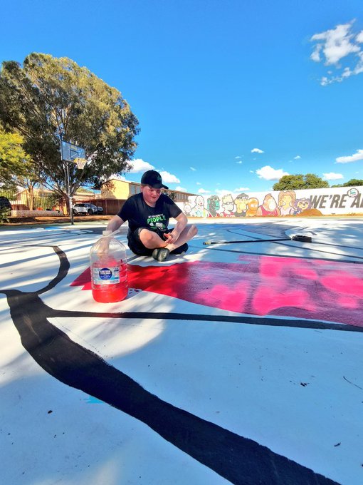 A man wearing a black cap and t-shirt kneels on an outdoor basketball court, painting a large red design next to a jug of red paint, with a basketball hoop and mural visible in the background.