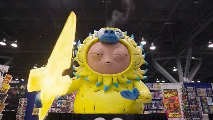 A large, yellow, furry collectible art toy figure with blue accents on its claws and face, displayed at what appears to be a convention, holding a large yellow lightning bolt shape.