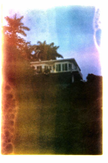 A vintage, blurry, hand-colored photograph of a white house on a hill surrounded by palm trees and dark foliage, with a strip of film perforations visible on the left edge.