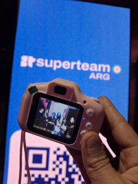 A hand holds a pink children's digital camera in front of a 'superteam ARG' sign and a QR code.