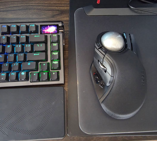An ergonomic trackball mouse rests on a black mousepad next to a backlit mechanical keyboard, complementing the post's joke about its "powerful" performance coinciding with a 10% rise in ETH.