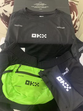 A black and neon green sportswear set from OKX (a cryptocurrency exchange), including a shirt branded "TRUSTED," "VERIFIED," and a fanny pack with "OKX" displayed in reflective text, is laid out on a bed—matching the post's excitement about receiving first-time OKX merchandise as a Mid-Autumn surprise.