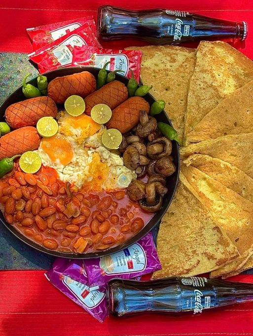 A traditional Mexican breakfast spread includes a skillet of eggs, sausages, beans, mushrooms, and limes, served with folded tortillas, Coca-Cola bottles, and ketchup and hot sauce packets on a red table.