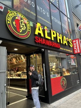A street-level view of an 'ALPHA'S SHAWARMA' restaurant, featuring yellow and red signage, with a person in a black hoodie standing near the entrance.