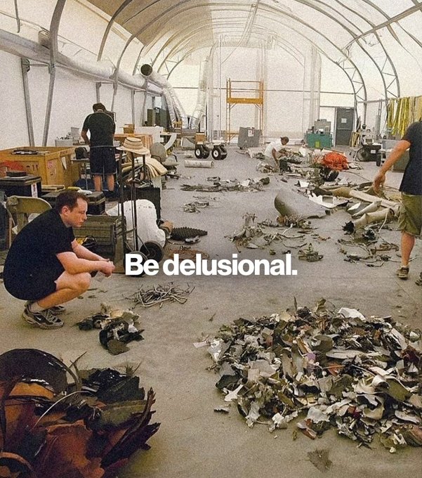 A group of people work inside a large tent-like structure surrounded by piles of scrap metal, debris, and industrial equipment, with the bold white text "Be delusional." overlaid in the center. The caption ties to "GM" by encouraging the automaker to embrace ambitious, unconventional thinking—suggesting bold vision is needed despite the chaotic, disassembled environment depicted.
