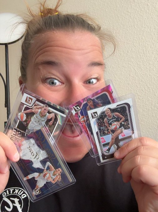 A woman with wide eyes holding three basketball trading cards in plastic sleeves in front of her face.