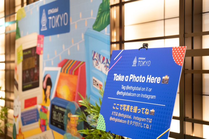 A sign at an ETHGlobal TOKYO event invites attendees to 'Take a Photo Here' and tag @ethglobal on X or Instagram, with bilingual English and Japanese instructions.