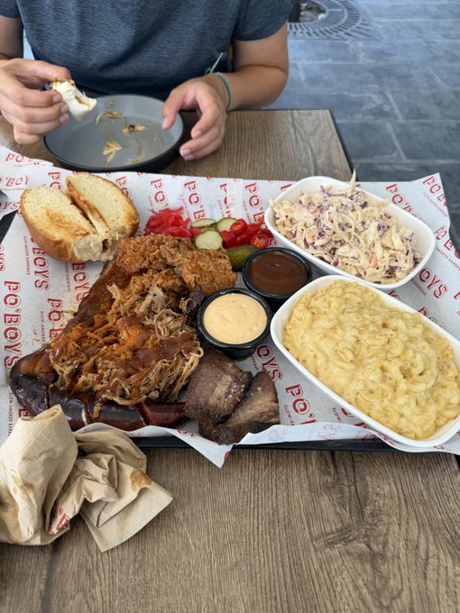 A BBQ feast from Po'Boys restaurant featuring pulled pork, brisket, sliced meat, coleslaw, mac and cheese, pickles, and bread, with a person eating at the table.