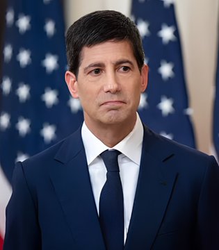 Portrait photograph of a man in a dark blue suit and tie standing in front of American flags.