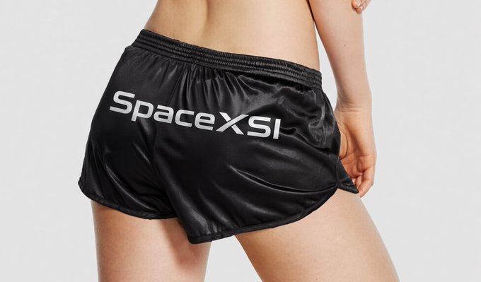 A close-up product shot of black satin shorts worn by a model, featuring the white "SpaceX" logo printed across the back.