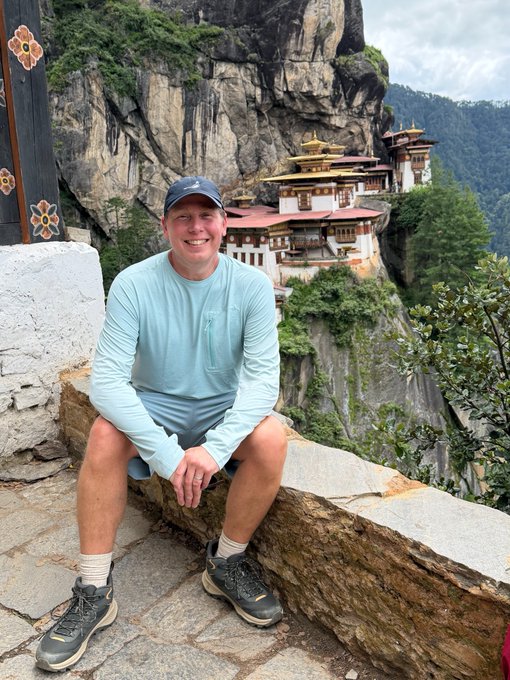 A smiling man in a light blue long-sleeve shirt, shorts, and a baseball cap sits on a stone ledge with the iconic Paro Taktsang (Tiger's Nest) Monastery dramatically perched on a cliff in the background, capturing a memorable moment of global exploration with his companion.