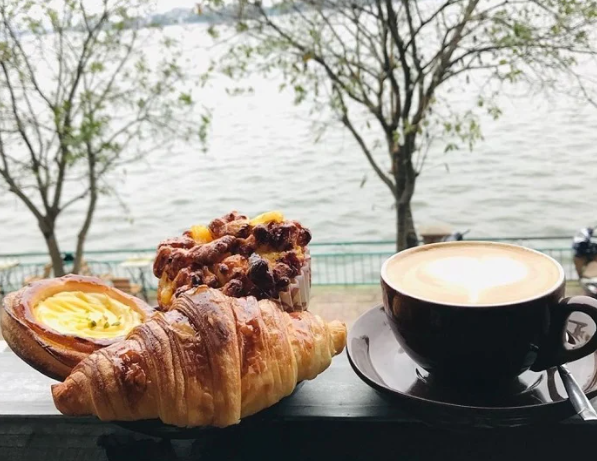 A close-up of a café table with a flaky croissant, a pastry, and a cappuccino with latte art, set against a scenic waterfront view with trees.