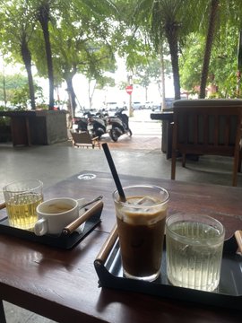 A wooden table at an outdoor café with two drinks (an iced coffee and a glass of water) on trays, with motorcycles and trees visible in the background.