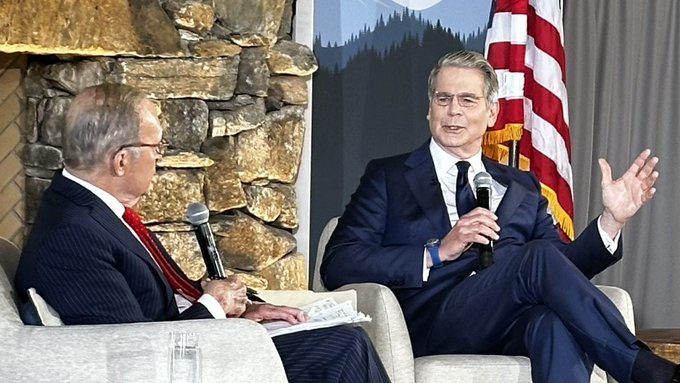 Two men in suits sit on a stage during an interview; the man on the right, wearing glasses and a blue suit, gestures while speaking into a microphone, with an American flag in the background.