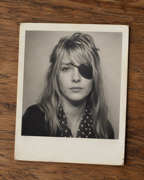 A vintage Polaroid-style black and white photo of a woman with long blonde hair wearing an eye patch over her left eye.