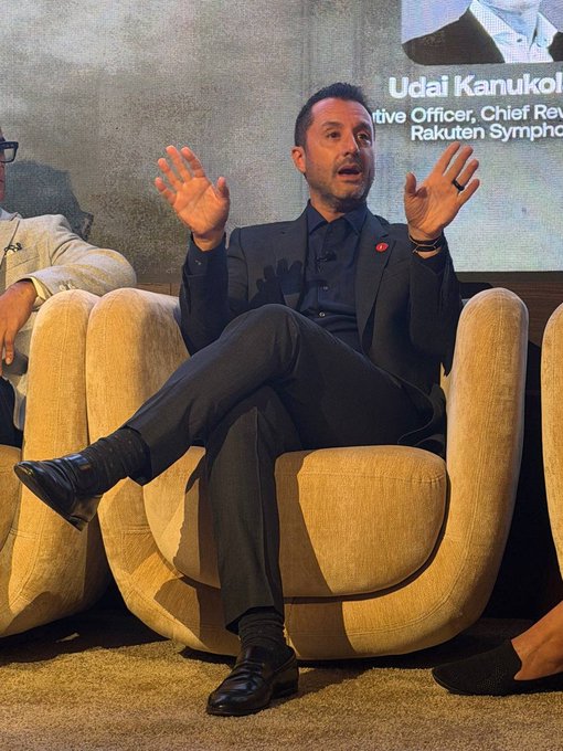 A man in a dark suit sits in a cream-colored armchair during what appears to be a panel discussion, gesturing expressively with both hands raised, with a screen behind him displaying text including "Udai Kanukol" and "Rakuten Sympho." The image complements the accompanying quote by visually capturing a keynote or panel moment where industry leaders, like John Nahas of AvaLabs, discuss bringing real businesses onchain through initiatives such as Iris on the Avalanche network.