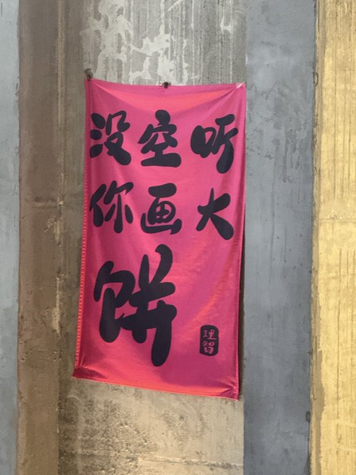 A bright pink banner with bold black Chinese calligraphy hangs against a weathered gray wall, prominently displaying the characters "没空呀 你画大饼" (roughly "No time, you're drawing big pies/empty promises"), with a small red seal reading "建智" at the bottom. The banner's message about empty promises visually echoes the post's skepticism about market timing and oversold investor sentiment following positive developments.
