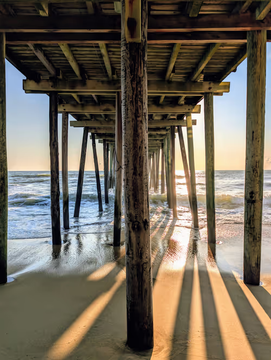 A view from underneath a wooden pier extending into the ocean, with sunlight filtering through the pilings and casting long shadows on the sandy beach during what appears to be sunset or sunrise.