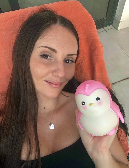 A woman with long brown hair poses on an orange towel holding a cute pink and white penguin squishy toy.