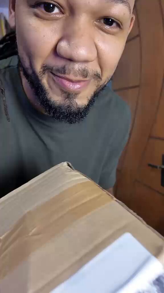 The image features a man holding an open cardboard box with a white label attached to it. The man has a beard and appears to be smiling at the camera, possibly posing for a photo or video. The background of the image is blurred, drawing attention to the man and his actions in front of him.