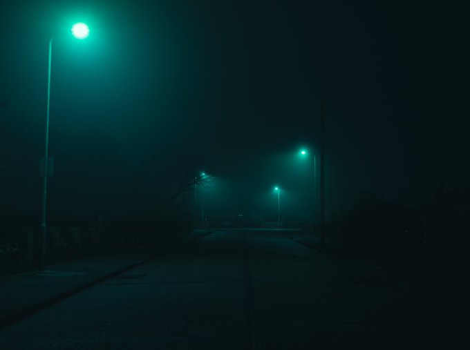 A dark, foggy street at night illuminated by cyan streetlights, creating an eerie and atmospheric scene.