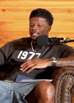 A young man wearing a "1977" black t-shirt sits in a podcast setting, speaking into a microphone while holding a notepad on his lap. The post discusses Houston Texans Hall of Famer Andre Johnson criticizing CJ Stroud's body language, suggesting the man in the image may be the featured speaker in the podcast clip.