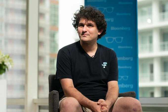 A photo of Sam Bankman-Fried sitting in a chair wearing a black FTX t-shirt during what appears to be a Bloomberg interview, with a cityscape visible through windows in the background.