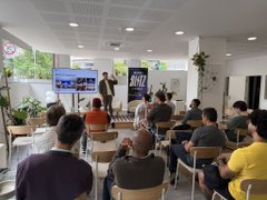 The image shows a group of people sitting in chairs around a table, watching a man giving a presentation using a large screen TV as a projection tool. The room appears to be a conference or meeting space with potted plants placed throughout the area. A sign is visible on one side of the room that reads "@LunarStrategy". The group seems engaged in the presentation and possibly discussing strategies related to the Monad Hackathon, which has attracted a large audience.
