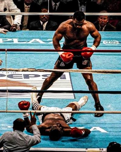 A famous boxing match photograph showing a boxer standing victorious with red gloves over his fallen opponent on the blue ring canvas, with spectators visible at ringside.
