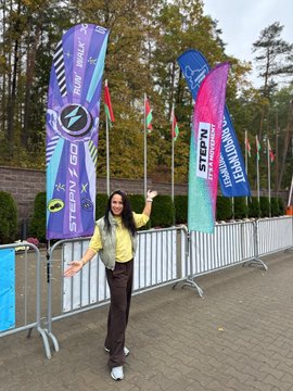 A woman in a yellow shirt and brown pants stands smiling with her arms outstretched in front of colorful event banners and flags, including one for 'STARDUST' and another reading 'WELCOME STREAMER'.