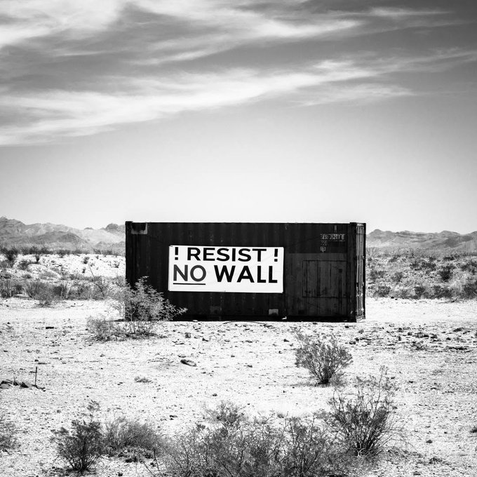 A black shipping container in the desert bears a sign reading "!RESIST! NO WALL," photographed near Terlingua, Texas—a fitting visual for a protest-themed NFT collection about opposing border wall construction.
