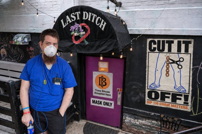 A masked man in a blue shirt stands outside a bar called "LAST DITCH" which displays signs for "BITCOIN Accepted Here," "MASK ONLY," and a "CUT IT OFF! RIP TIO CHAIN MATTERS" poster.