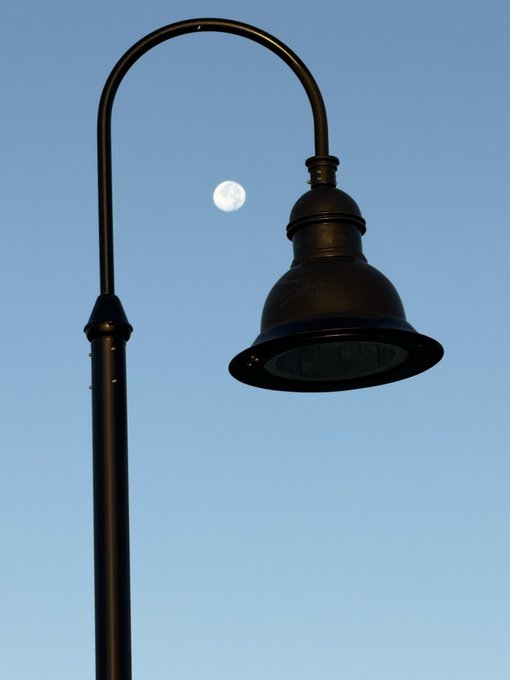 A black vintage-style street lamp silhouetted against a clear blue sky with the full moon visible through its curved frame.