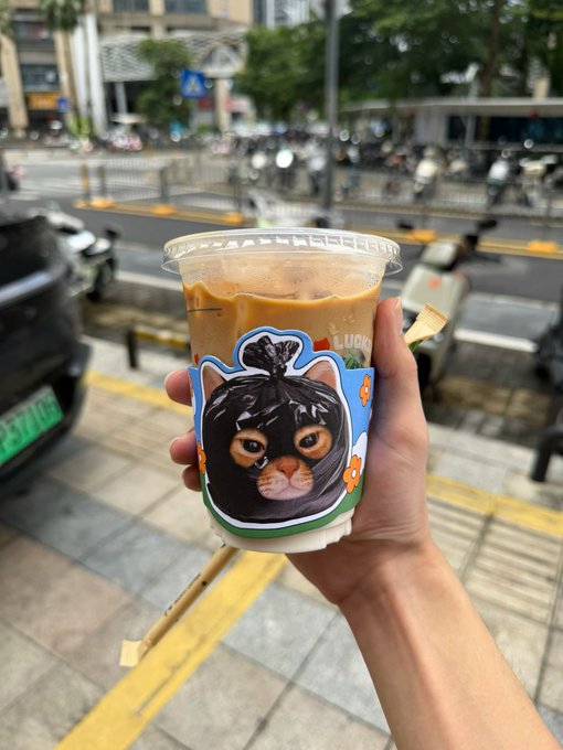 A hand displays a plastic cup of iced coffee featuring a decorative cardboard sleeve with an illustration of a black cat, set against a blurred urban street background with parked scooters and a green license plate reading "B71061" visible on the left.