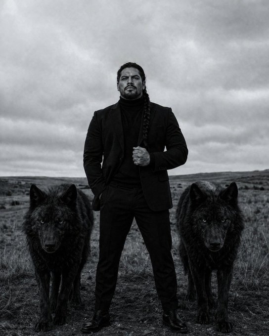 A man in a black suit stands confidently between two black wolves on an overcast day, embodying the post's message about resilience and not underestimating strength. The stoic figure flanked by fierce wolves visually reinforces the quote about recognizing true power that may appear softened by hardships.